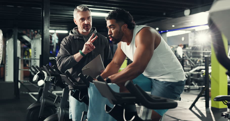 Coaching, cycling or time with men in gym for fitness machine, wellness or cardio workout. Stationary bike, exercise or personal trainer with intensity in sports club for smart watch and health stats