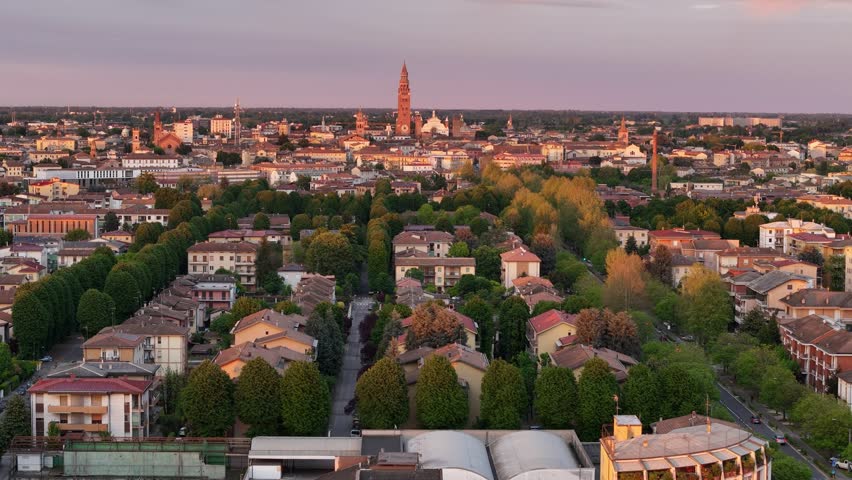 Aerial pull-back view over Cremona, Lombardy, Italy, showcasing tree-lined avenues and residential buildings with the iconic Torrazzo di Cremona and cathedral bathed in golden hour sunlight.