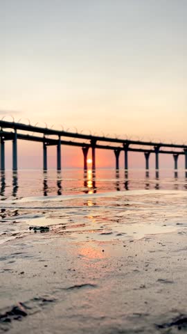 Beautiful sunset over the sea. The sun setting between the pillars of the bridge. Reflection of sunlight on water. Kanonersky Island, Saint-Petersburg. Vertical FullHD slow motion footage.