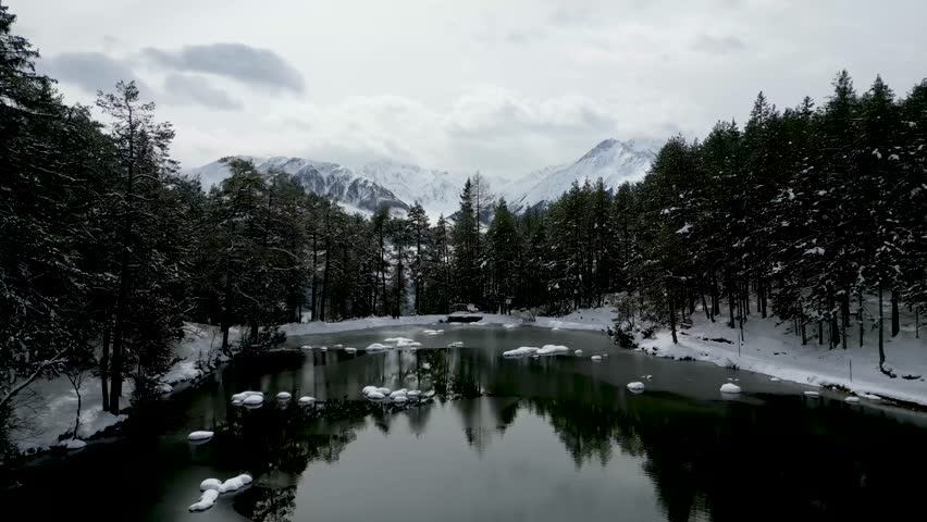 Serene Winter Landscape: Drone Flight Over a Partially Frozen Lake and Snow-Capped Mountains with a Picturesque Village Below