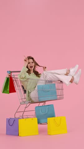 A cheerful young woman sits in a shopping cart, surrounded by colorful shopping bags, in a vibrant setting.