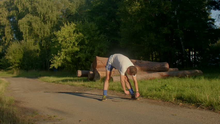 A man performs a stretching routine by a quiet road, kneeling by logs to warm up his legs, all in a serene outdoor setting