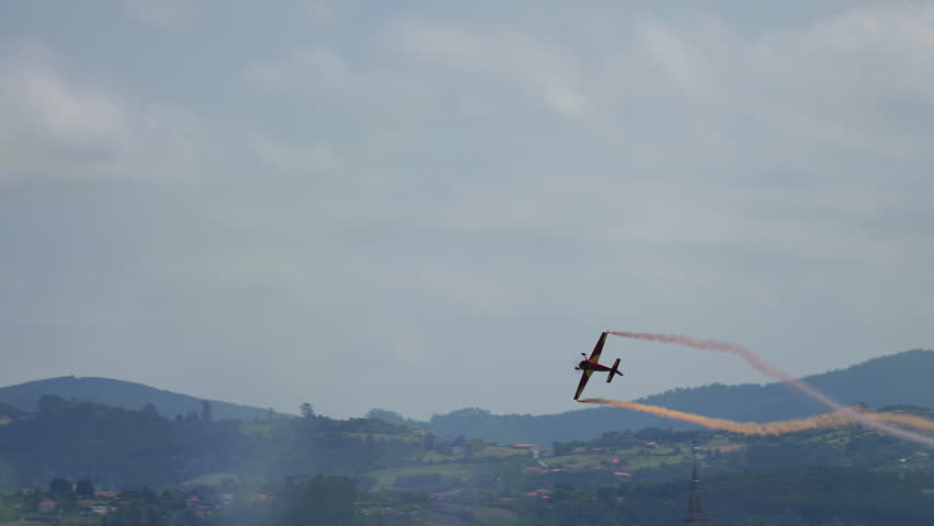 Single airplane with orange smoke trail performing aerial stunt above green hills during airshow