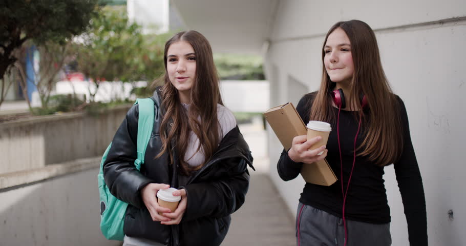 Two Teenage Girls Walk and Talk Together Between Classes at School While Holding Takeaway Coffee Cups