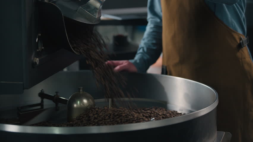 Roasted coffee beans fall out roaster machine hot fresh steam aroma caffeine roasting unrecognizable man guy check quality barista business owner industry working in factory blend roast cropped view