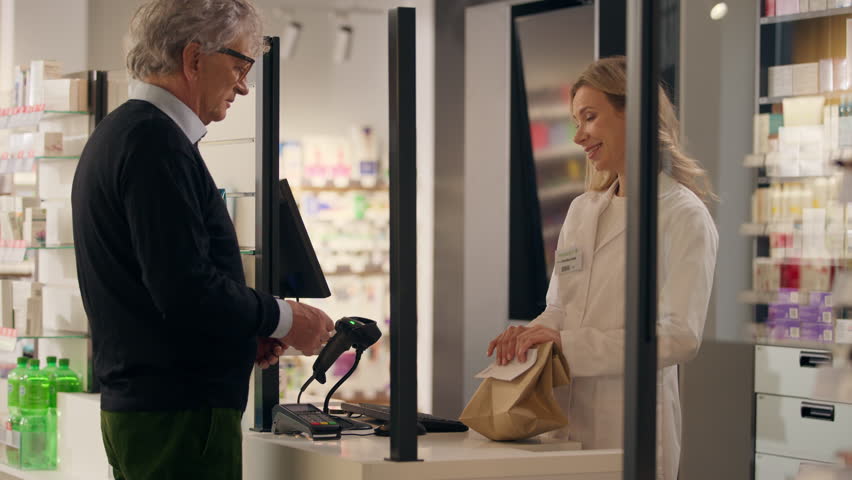 Pharmacy professional medical help smiling friendly woman pharmacist consultant selling medicine at cashier counter old man customer buying medication in paper bag drugs pills health order drugstore