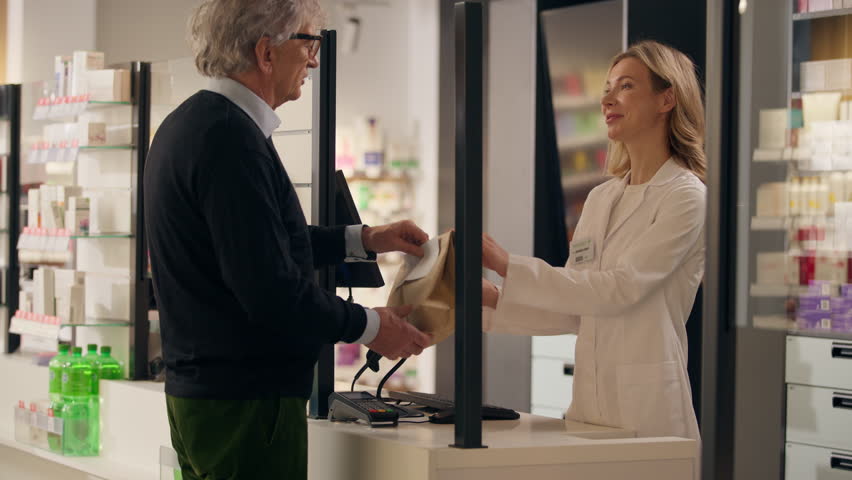 Pharmacy professional medical help smiling friendly woman pharmacist consultant selling medicine at cashier counter old man customer buying medication in paper bag drugs pills health order drugstore