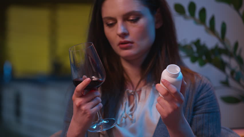 Sad upset woman holding glass wine pills thinking difficult choice sick unwell drink alcohol addiction worried girl drinking antibiotic health problem disease frustrated Caucasian female at home night