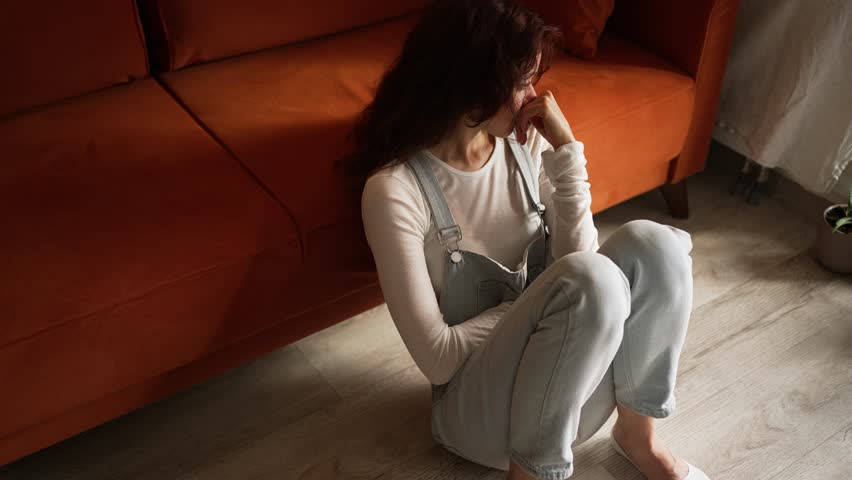 Sad woman sitting on the floor by an orange sofa at home, suffering from depression, loneliness, headache or other mental health issues, crying and covering her face