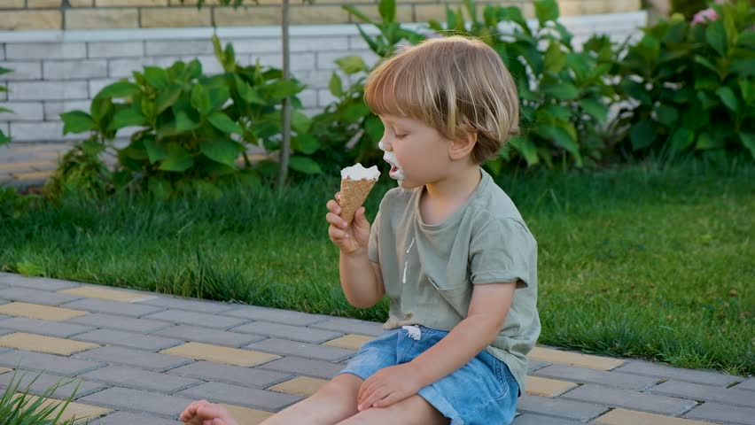 Cute toddler child eating ice-cream cone sitting on grass outside. Kids summer fun joy, holiday, vacation on sunset light. Dirty mouth, happy childhood.
