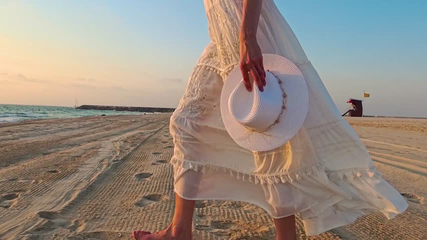 Woman in white summer maxi dress walking into the water on beach holding white hat in hand at sunset. Kite beach, Dubai