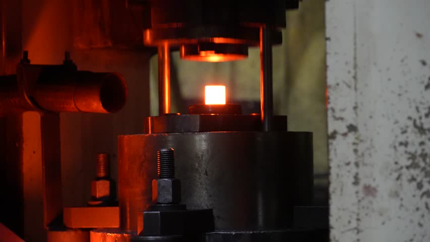 Glowing hot metal piece secured on a fixture with threaded rods and bolts. High-temperature industrial process used in forging, heat treatment, or precision metalworking under dim lighting.