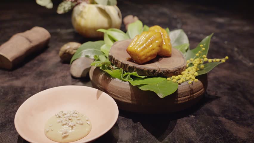 Pan zoom of madeleines placed on a rustic wooden pedestal adorned with native leaves and yellow acacia blossoms. Paired with a smooth native sauce served in a soft pink bowl.