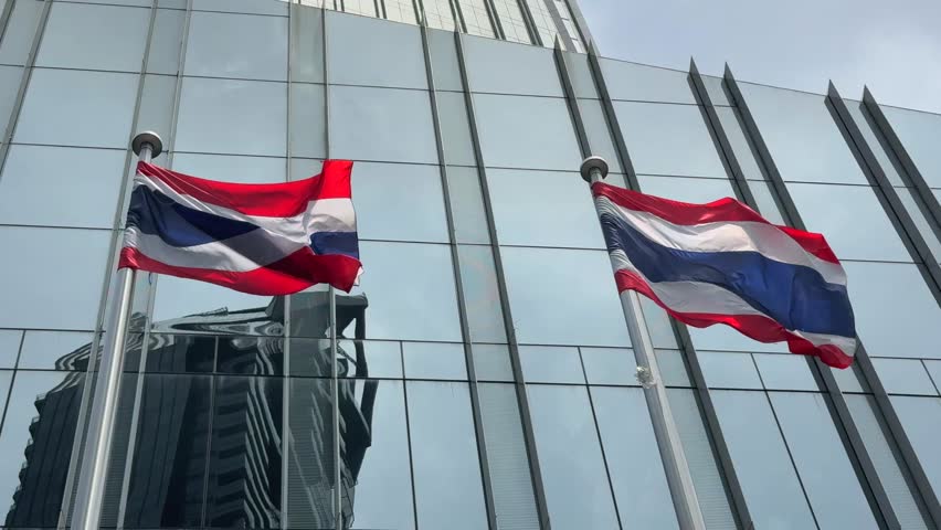Slow motion of Two Thai flags waving in the wind in front of a mirror-glass building in Bangkok. Urban city life and modern architecture meet national pride and tradition.