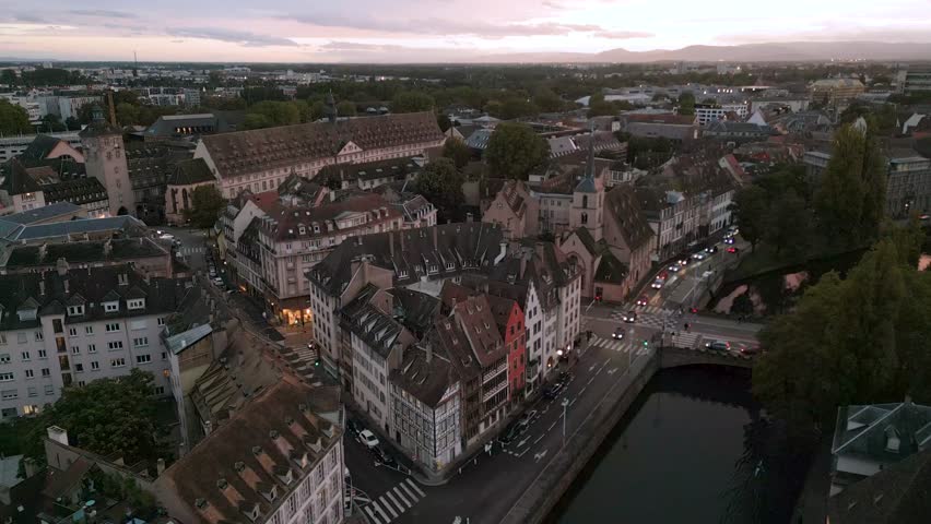 Atmospheric twilight aerial view over Strasbourg’s winding streets and rooftops, with soft evening lights illuminating the historic architecture and calm riverbanks as the city transitions into night