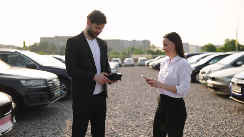A male car dealer hands the keys to a woman who is buying a car on a sunny day.