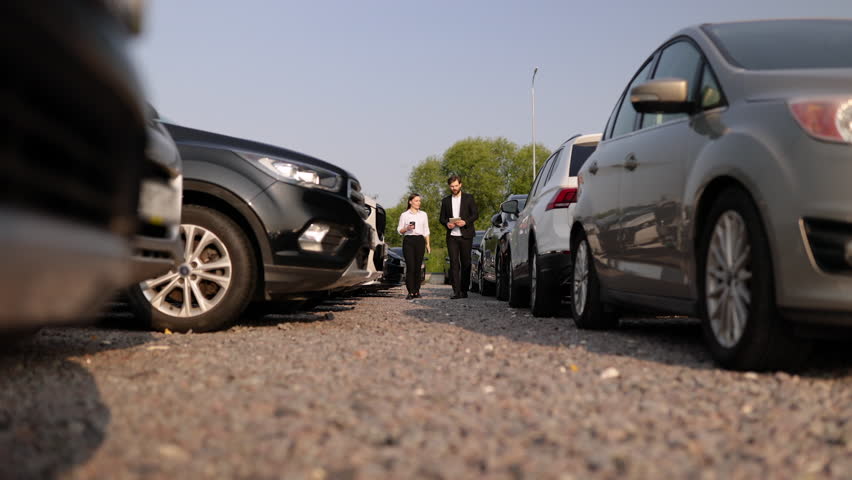A male car dealer showing a car to a woman on a car lot, with a row of vehicles in the background.