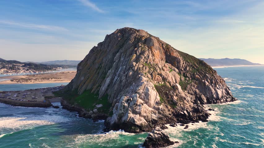 Drone establishing orbit of Morro Rock and coastal waves in Morro Bay, California under sunny skies