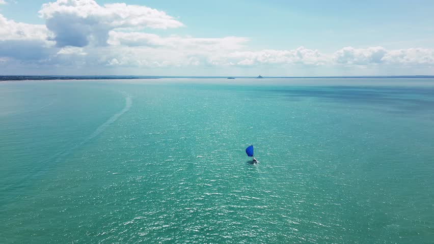 Drone footage captures a solitary sailboat gliding peacefully across the water toward Mont Saint-Michel, visible in the distant horizon.