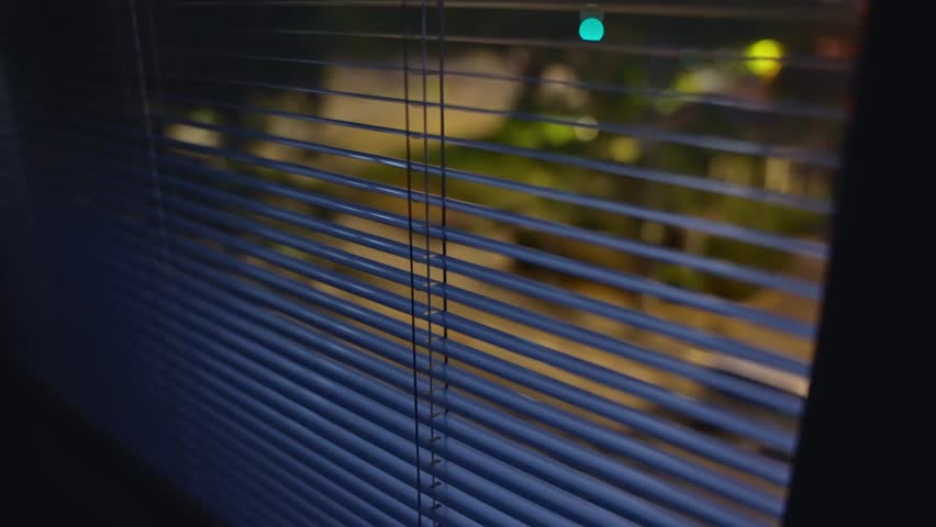 Close up on window shutters or blinds at night overlooking quiet city street. Suspicious person point of view pov shot. Hidden spying or searching for something outside. Secret operation or hideout