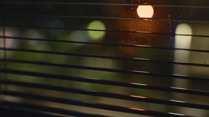 Window shutter blinds at night overlooking city street. Defocused street lights shining. Secret hidden view. Person searching or spying during dark hours. Police or covert operation investigation