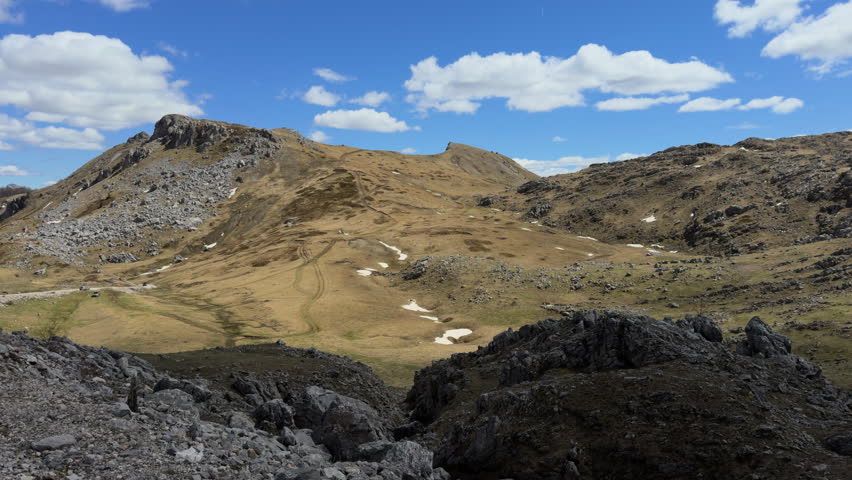 A scenic mountain-top view captured from a moving car, showcasing a rugged landscape of rocky terrain and low grass-covered fields with narrow twisted road. slow motion, sunny day, wide shot