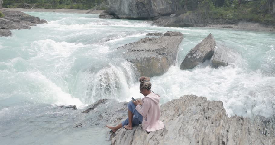 Black lady on her phone sitting on the rocks near the edge of a waterfall (Natural Bridge lower falls) and beautiful scenic blue river in Yoho National Park.