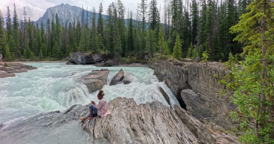 Black lady on her phone sitting on the rocks near the edge of a waterfall (Natural Bridge lower falls) and beautiful scenic blue river in Yoho National Park.