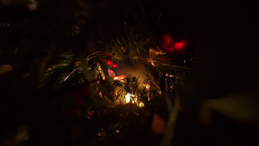 Hand Hanging Red Ornament Among Pine Branches with Glowing Lights and Ribbons in Zoomed View Representing Holiday Tree Decoration Tradition.