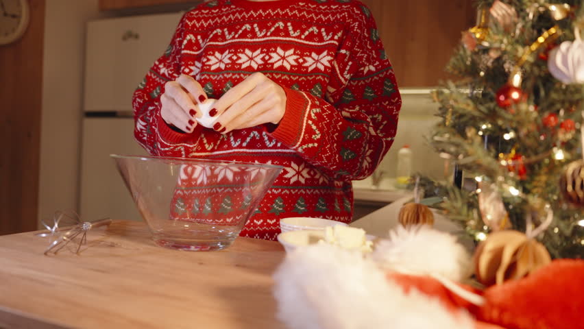 Woman in Red Christmas Sweater Cracking Egg into Glass Bowl on Wooden Table While Preparing Dough in Warm Kitchen, Holiday Baking Mood.