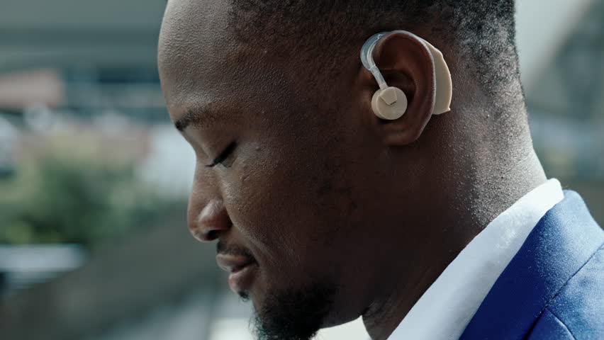 Closeup portrait of African American office employee with hearing aid on urban backdrop. Young businessman with disability shows diversity and corporate culture values on street