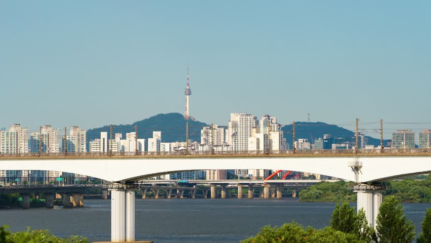 A subway trains travel across the Dangsan Railway Bridge in both directions over the Hangang River, with the iconic Namsan Tower and the sprawling cityscape of Seoul, South Korea, in the background.
