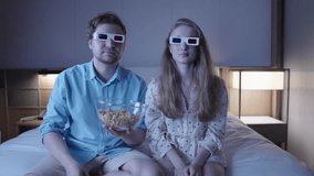Happy young couple wearing 3d glasses are sitting on a bed at home, watching a movie and eating popcorn, reacting with surprise and excitement to the film, in slow motion - Powered by Shutterstock - Get 15% off with code: PIKWIZARD15