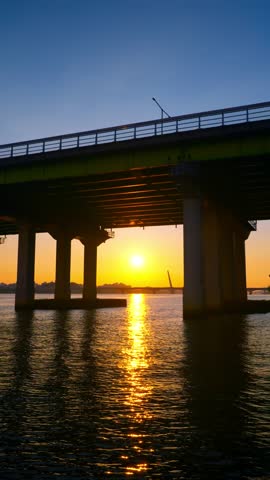 A beautiful vertical shot from the riverfront shows the golden sun setting over the Hangang River, framed by the silhouette of the Yanghwa Bridge in Seoul, South Korea.