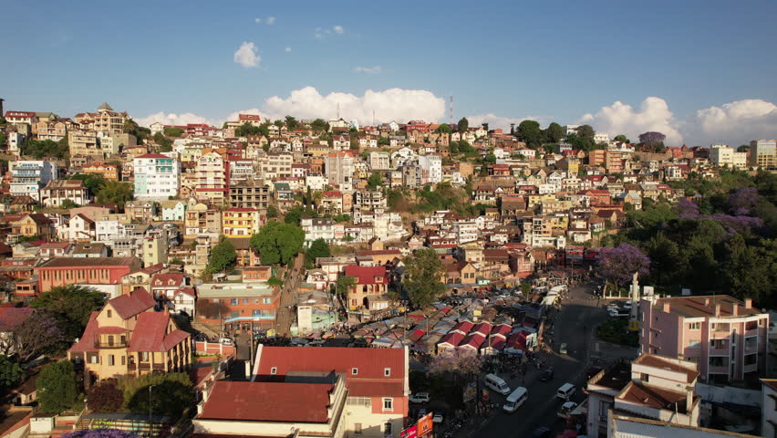 A short aerial clip revealing the edge of Ambohijatovo Market in Antananarivo, Madagascar, with jacaranda trees blooming in the lower frame and layers of hillside homes above.