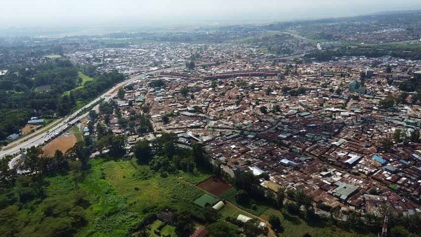 High angle shot of Nairobi, Kenya reveals a densely populated urban slum adjacent to green areas. A highway cuts through both the urban sprawl and verdant landscape.