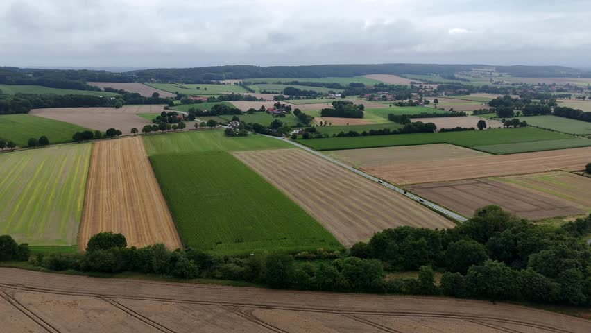 Aerial view of rural farmland in American Midwest. Patchwork fields, country roads and scattered farmhouses under a cloudy sky. Summer day with cars on rural intersection road. Wide shot.