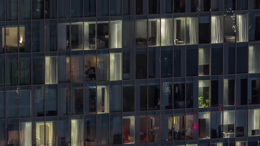 Frontal view of night facade of tower building with a lot of windows in apartments with light timelapse. Life in every window. Aerial view of the glass facade of an office and residential skyscraper.
