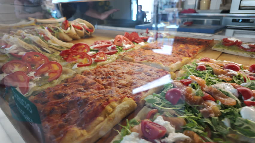 Italian bakery display with focaccia and pizza al taglio topped with tomatoes, pesto, and mozzarella, highlighting traditional street food and savory pastries