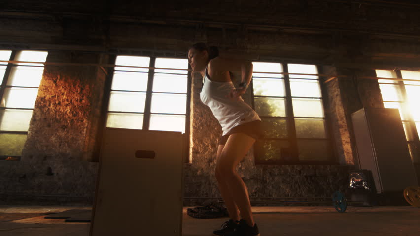 Fit Woman Performs Box Jumps in an Industrial Gym With Stone Walls, Large Windows, and Sunlight Streaming In, Focusing on Lower Body Strength, Agility, Cardio Exercise.