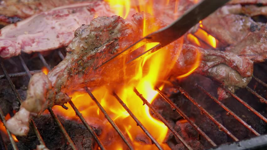A chef uses tongs to flip a sizzling lamb steak on a hot barbecue grill. Bright flames flare up, searing the meat and creating a perfect crust. The ultimate grilling moment.