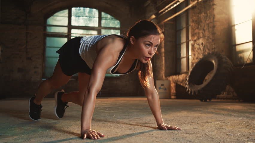 Athletic Woman With Long Brown Hair in a Ponytail Performs Mountain Climbers on the Gym Floor, Wearing a Tank Top and Black Shorts, in an Industrial Gym