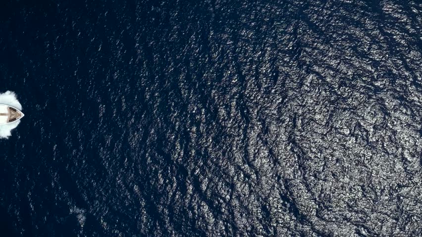 Aerial overhead view of a luxury yacht passing by over blue, sparkling ocean leaving a trail of white surf and waves