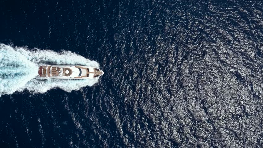 Aerial overhead view of a luxury yacht passing by over blue, sparkling ocean leaving a trail of white surf and waves