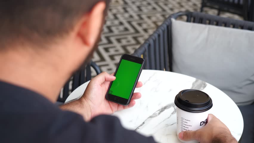 Man sitting at a café table holding a cardboard coffee cup and looking at a green screen smartphone. The cup is logo-free and the phone screen is editable—ideal for branding or mockups