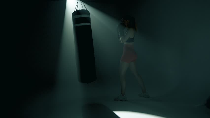 Blonde fit woman throws strong jabs and hooks at a heavy bag using both hands. Full body shot with slow camera push-in. Powerful, energetic punches in smoky gym with dramatic cinematic lighting