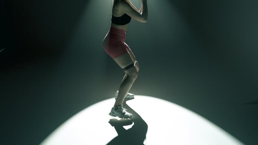 Blonde woman in gym performs side lunges with resistance band above knees. Cinematic light creates glowing rim on her body. Full body tracking shot from feet to head in smoky dark training studio