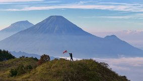 Majestic mountain landscape with a person waving a red - white (Indonesia) flag on a grassy hilltop above the clouds. The concept of pride in one's nation and state. Aerial drone view. - Powered by Shutterstock - Get 15% off with code: PIKWIZARD15