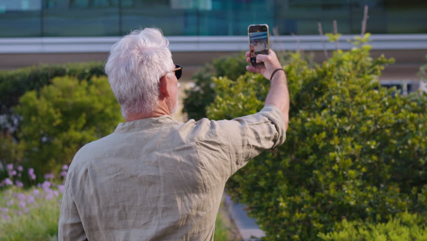 Senior businessman wearing sunglasses taking selfie with smartphone while turning around to look at camera. He stands outdoors in garden area with greenery in daylight