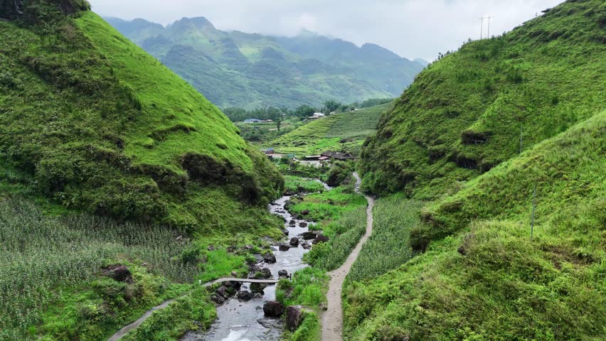 A rocky stream winds through lush green hills with a narrow path beside it, leading to a distant village in Ha Giang
